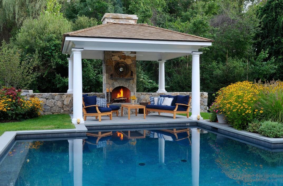 Un gazebo barbecue in stile classico si trova sullo sfondo di alberi e fiori decorativi, che si riflette sulla superficie dell'acqua della piscina